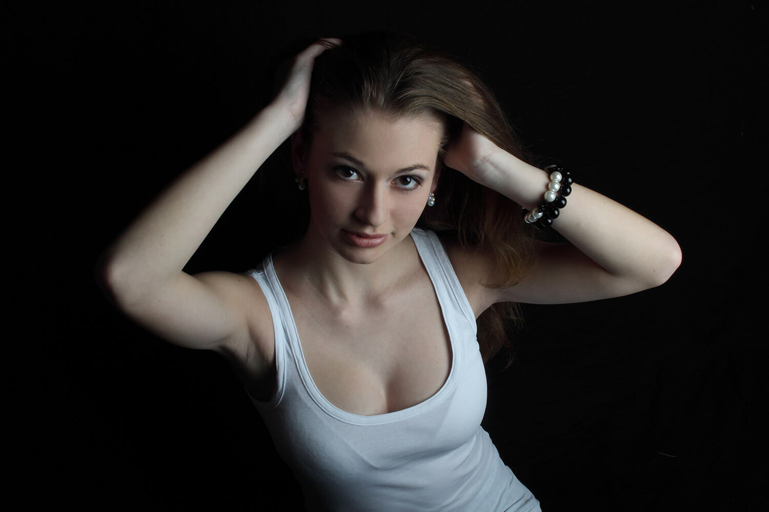 female portrait in a white tank top and with her hands in her hair on a dark background